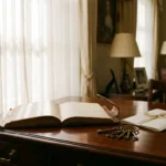 Leather-bound ledger open on a mahogany desk beside antique house keys and a folded document in a traditional British sitting room.