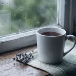 White ceramic mug of steaming tea on a weathered windowsill with rain-streaked glass, dried lavender sprig alongside in soft grey British daylight.